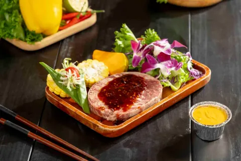 Tảng bò nướng phủ sốt phô mai trứng muối trên thớt gỗ, kèm bắp, hoa lan và chén sốt